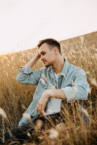 Young handsome man, wearing denim jacket, sitting in a field, looking to side. © Bostan Natalia