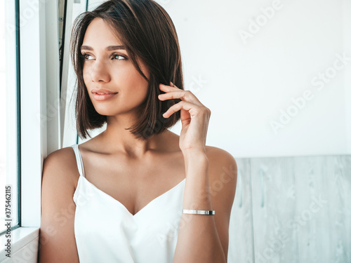 Portrait of beautiful woman dressed in white pajamas. Sexy carefree model enjoying her morning at balcony. She dreams about something and looking out the window. Female in her thoughts