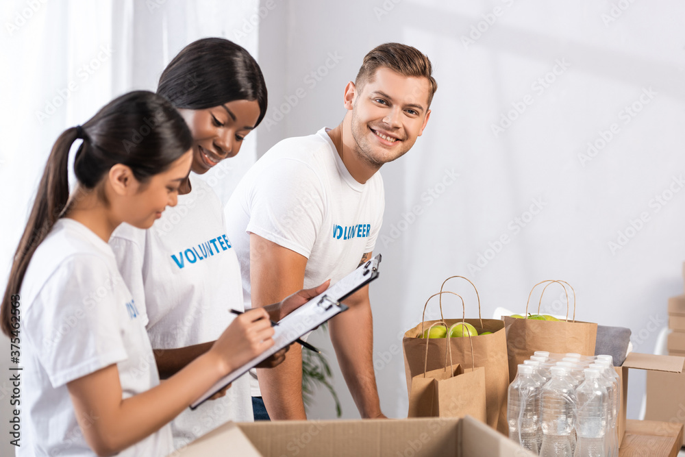 Selective focus of volunteer looking at camera near multicultural women with clipboards and packages in charity center Selective focus of volunteer looking at camera near multicultural women with clipboards and packages in charity center