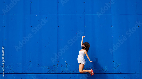 Woman with medical mask and white dress jumping over a blue wall