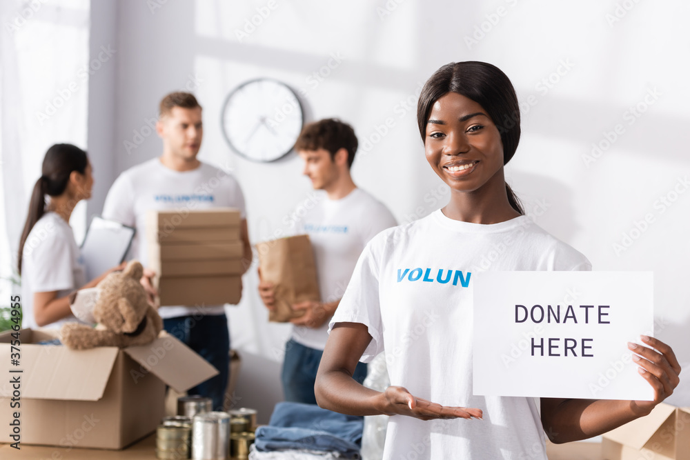 Selective focus of african american volunteer pointing at card with donate here lettering in charity center Selective focus of african american volunteer pointing at card with donate here lettering in charity center