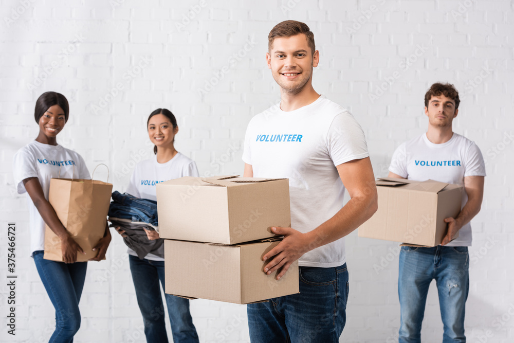 Selective focus of man holding carton boxes near volunteers with paper bag and clothes at background Selective focus of man holding carton boxes near volunteers with paper bag and clothes at background