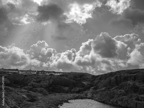 Rocky landscape on Swedish coast. Part of the ocean is in the foreground and houses are on top of the cliffs. It is a monochrome photograph with nice clouds and sunlight.