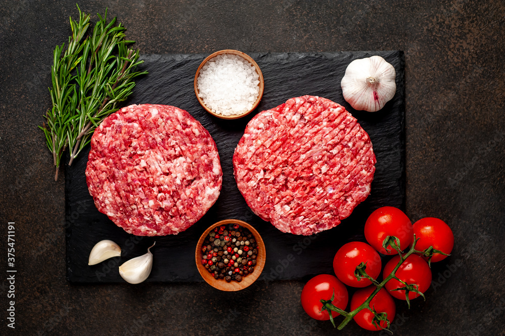 Raw beef burger cutlets with rosemary and spices on a slate board on a concrete background