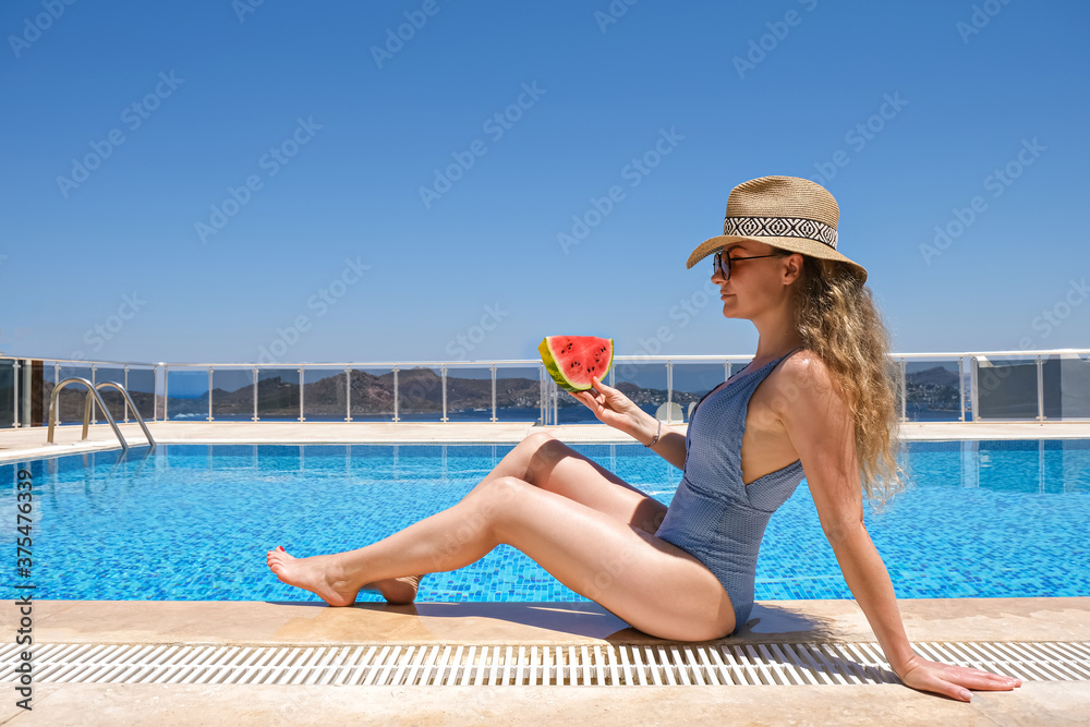 Young woman with slice of watermelon in straw hat sunbathing sitting on the edge of swimming pool