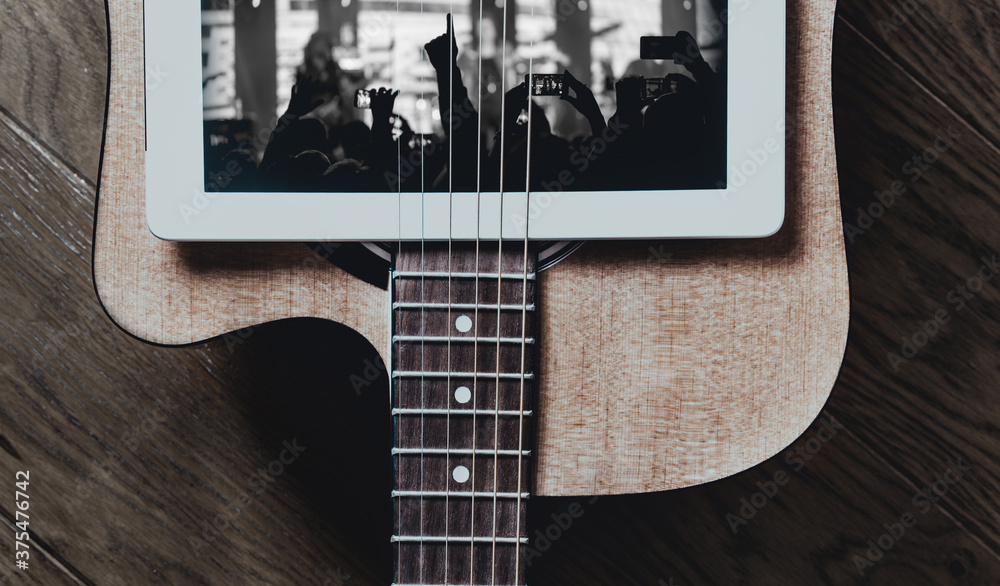 Fototapeta premium Concept of new normal in music. Combination of an acoustic guitar and a crowd of people on a tablet screen. Contrast the current insulation and fun time before a pandemic.