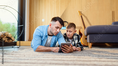 Young boy with father lying on the floor using tablet pc in modern apartment. Happy family time together.