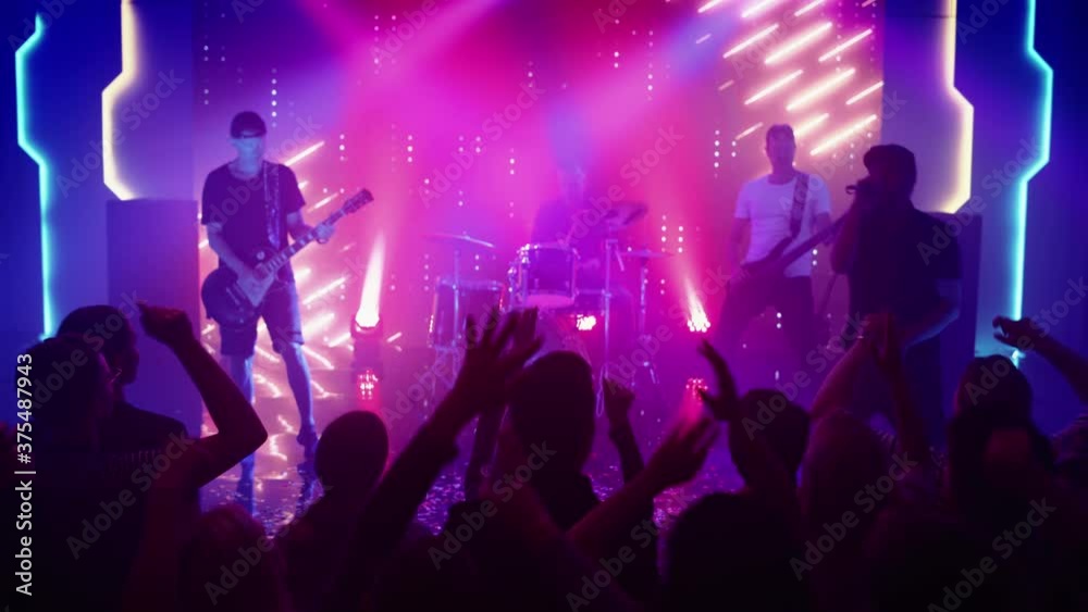 Rock Band with Guitarists and Drummer Performing at a Concert in a Night Club. Front Row Crowd is Partying. Silhouettes of Fans Raise Hands in Front of Bright Colorful Strobing Lights on Stage. 