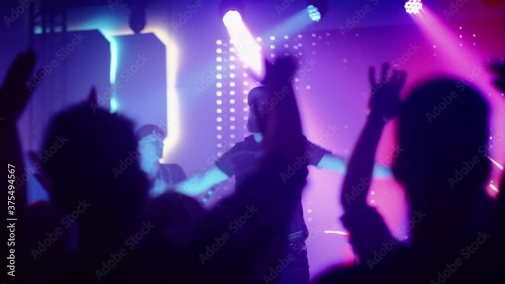 Rock Band with Guitarists and Drummer Performing at a Concert in a Night Club. Front Row Crowd is Partying. Silhouettes of Fans Raise Hands in Front of Bright Colorful Strobing Lights on Stage. Rock Band with Guitarists and Drummer Performing at a Concert in a Night Club. Front Row Crowd is Partying. Silhouettes of Fans Raise Hands in Front of Bright Colorful Strobing Lights on Stage.
