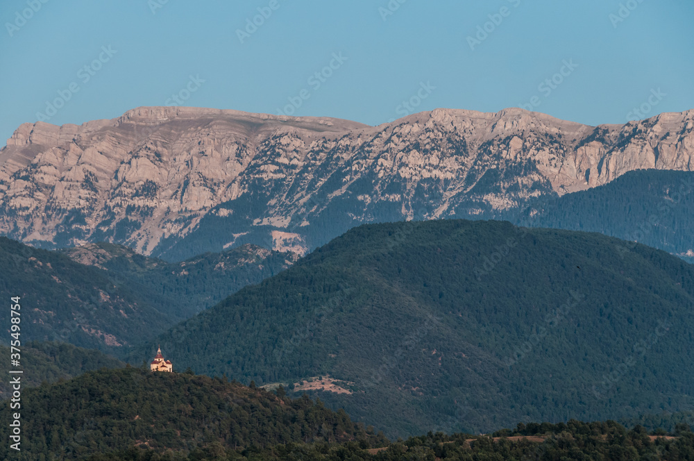 Obraz premium Pequeña capilla aislada en una cima frente a la imponente cordillera montañosa de los Pirineos en la provincia española de Lérida.