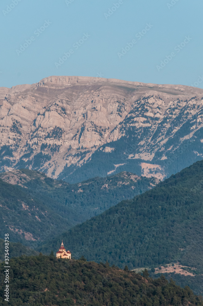 Obraz premium Pequeña capilla aislada en una cima frente a la imponente cordillera montañosa de los Pirineos en la provincia española de Lérida.