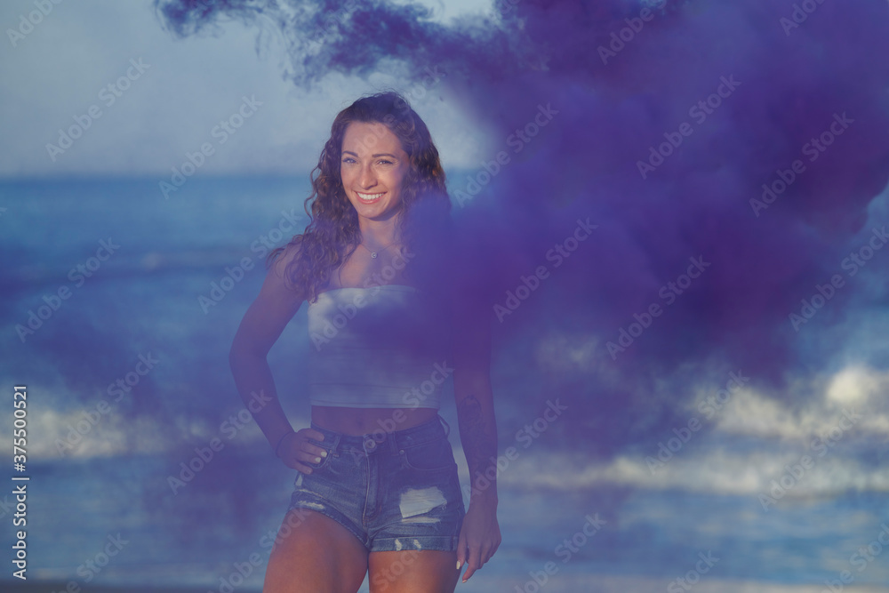 Obraz premium Beautiful young brunette woman in white shirt and denim shorts poses on beach - with purple smoke in air near her