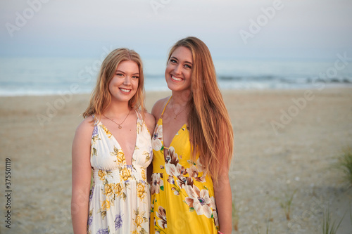 Two beautiful blonde sisters laugh as they pose together for family photo on the beach - standing in sand in dresses