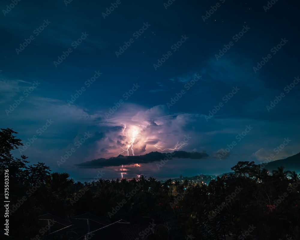 Lightnings, stars and dramatic clouds in the night sky over valley with trees, sea,  city and mountains. View from the top.