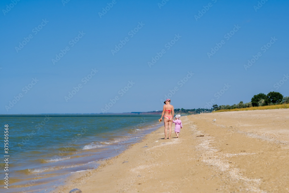 Rear view of a mother and a baby girl walking along a deserted sandy sea beach along the surf against a blue cloudless sky. A woman and a child are holding hands. Sunny day. Family vacation concept.