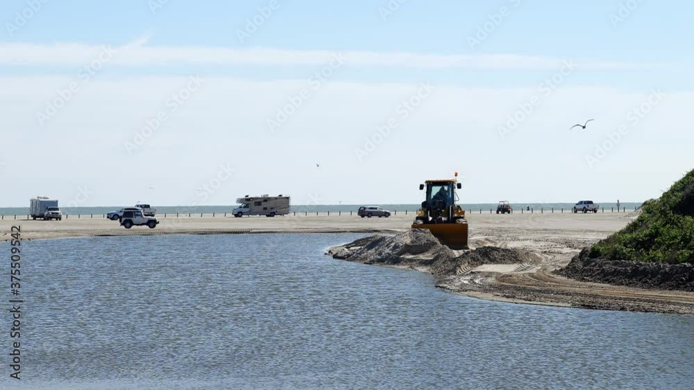 Beach scene in Port Aransas, Texas with vehicles parked and driving on the sand, and a front end loader moving sand near a holding pond. Gulf of Mexico is in the distance.
