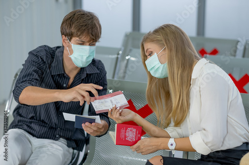 Couple Caucasian lovers man and woman with mask sit on chair with social distance and discuss about boarding pass in airport during flight for travel after relieve of lockdown from Covid-19 pandemic.