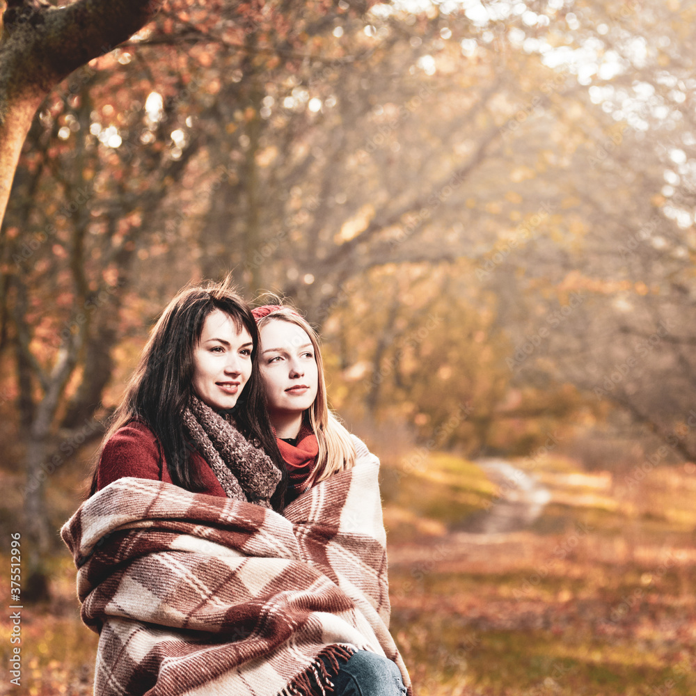 Pretty young woman covered with plaid with her attractive friend while walking in autumn park
