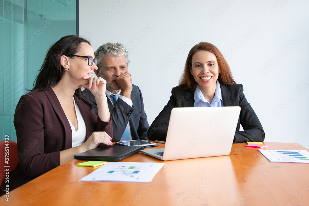 Happy business team watching project presentation on laptop, sitting at meeting table with paper reports and diagrams. Medium shot. Marketing and teamwork concept Happy business team watching project presentation on laptop, sitting at meeting table with paper reports and diagrams. Medium shot. Marketing and teamwork concept