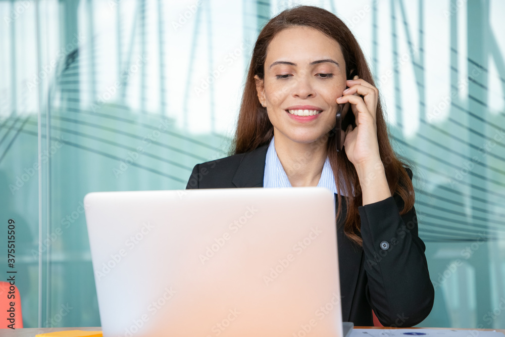 Positive pretty young businesswoman talking on mobile phone and smiling, working at computer in office, using laptop at table. Medium shot, front view. Communication or multitasking concept Positive pretty young businesswoman talking on mobile phone and smiling, working at computer in office, using laptop at table. Medium shot, front view. Communication or multitasking concept