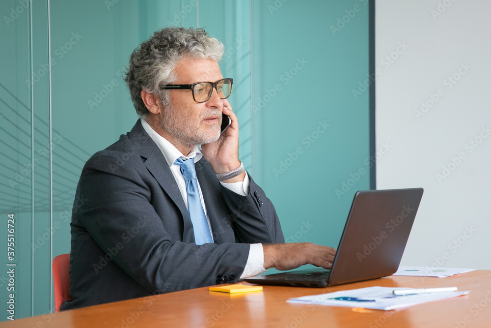 Serious mature executive in suit and glasses talking on mobile phone, working at computer in office, using laptop at table with paper diagrams. Medium shot. Communication or multitasking concept Serious mature executive in suit and glasses talking on mobile phone, working at computer in office, using laptop at table with paper diagrams. Medium shot. Communication or multitasking concept