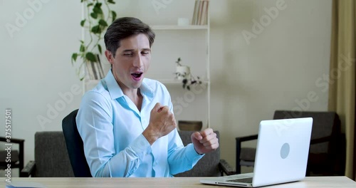 Young funny manager, businessman fooling around in office sitting at laptop, dancing in workplace, beating his heart rhythmically with hands in chest like drum, hums song, feels carelessness happiness