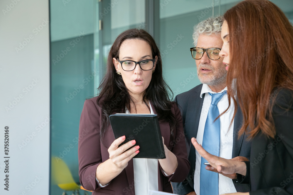 Surprised businesspeople looking on tablet screen and standing in office room. Brunette businesswoman in glasses holding digital device and showing data to colleagues. Business and teamwork concept Surprised businesspeople looking on tablet screen and standing in office room. Brunette businesswoman in glasses holding digital device and showing data to colleagues. Business and teamwork concept