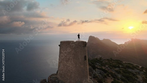 Aerial orbit shot of the silhouette of a man standing on top of an old watchtower at sunrise photo