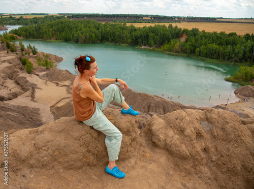 A woman with red hair and dark glasses sits on a high mountain above a bright blue lake .