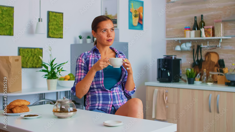 Lady enjoying a hot green tea sipping from porcelain cup. Woman drinking standing at the table in the kitchen and looking through the glass holding tea cup with natural, aromatic herbal drink. Lady enjoying a hot green tea sipping from porcelain cup. Woman drinking standing at the table in the kitchen and looking through the glass holding tea cup with natural, aromatic herbal drink.