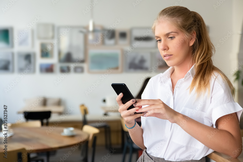 Focused beautiful blonde woman wearing white shirt, using smartphone standing in co-working space. Medium shot, copy space. Digital communication concept Focused beautiful blonde woman wearing white shirt, using smartphone standing in co-working space. Medium shot, copy space. Digital communication concept
