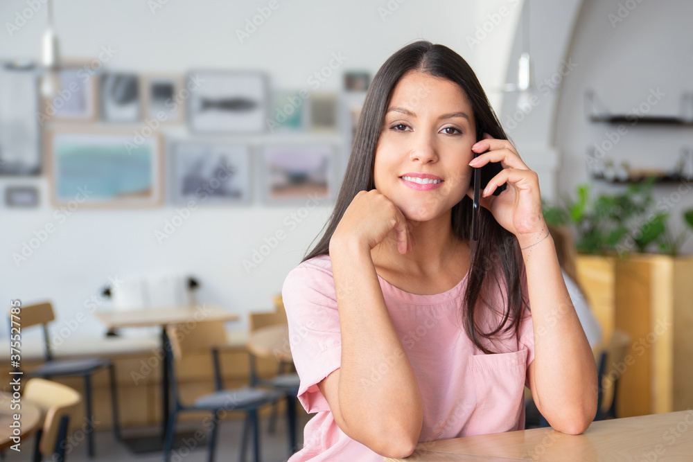 Fototapeta premium Happy confident beautiful young woman talking on cell, standing at co-working, leaning on desk, looking at camera and smiling. Medium shot, copy space. Digital communication concept