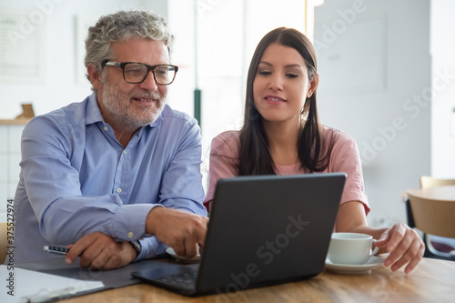 Pensive smiling mature man and young woman sitting at open laptop, looking and pointing at display, watching content over cup of coffee. Medium shot. Digital communication concept