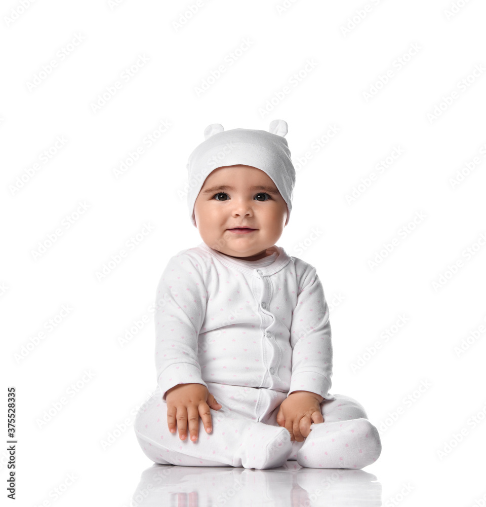 Infant baby toddler sits on the floor in white jumpsuit overall and funny hat with ears touching her legs Infant baby toddler sits on the floor in white jumpsuit overall and funny hat with ears touching her legs