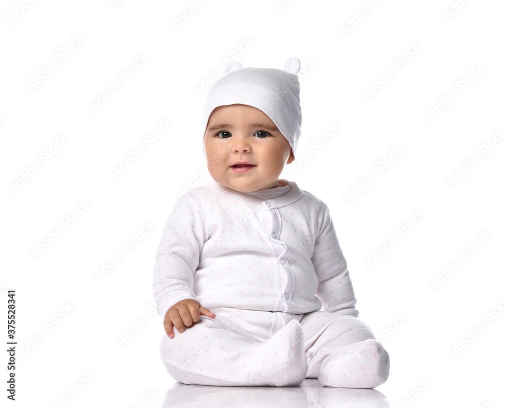 Attentive infant baby toddler in white jumpsuit overall and funny hat sits on the floor listens and tries to understand Attentive infant baby toddler in white jumpsuit overall and funny hat sits on the floor listens and tries to understand