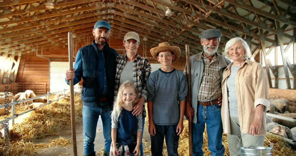 Portrait of happy Caucasian family of three generations standing in shed with livestock and smiling. Old parents with children and grandchildren in stable. Farmers with kids at farm. Zooming out.