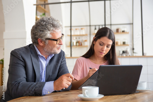 Mature professional and young customer signing agreement. Man and woman sitting at table with laptop and discussing papers. Expertise and paperwork concept