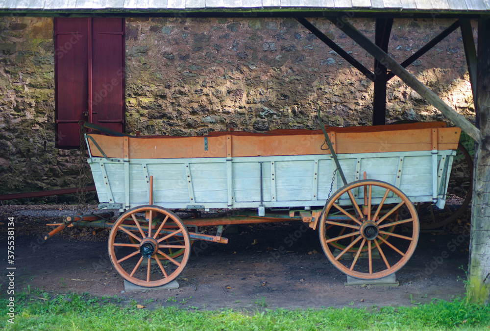 Early American wagon sits by a beautiful stone wall with red window shutters at historic village. Beautiful restored construction and color at Hopewell Furnace National Historic Site in Pennsylvania.