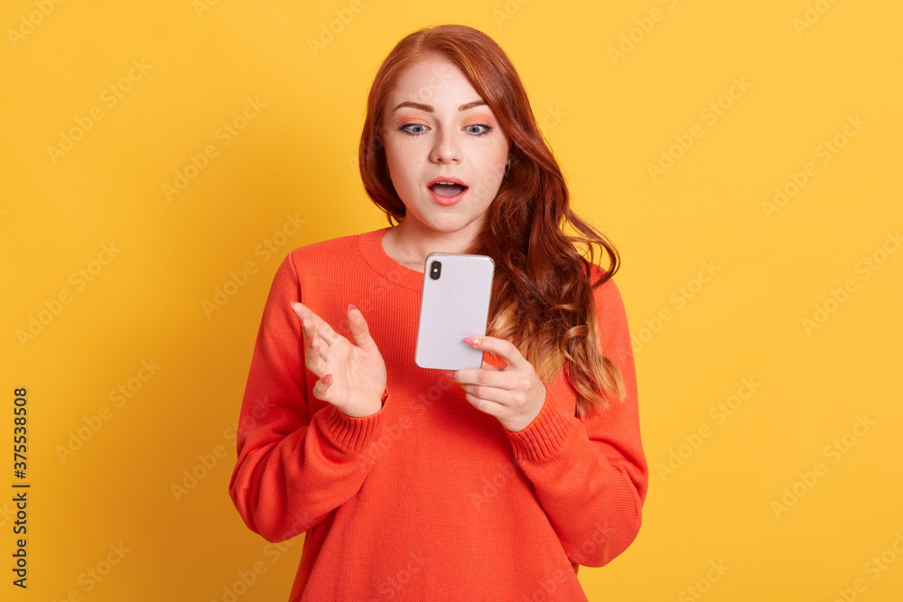 Stunned European woman being in shock, reads terrible news from cellular gadget, has wavy red hair, wears orange sweater, poses against yellow wall. Stunned European woman being in shock, reads terrible news from cellular gadget, has wavy red hair, wears orange sweater, poses against yellow wall.