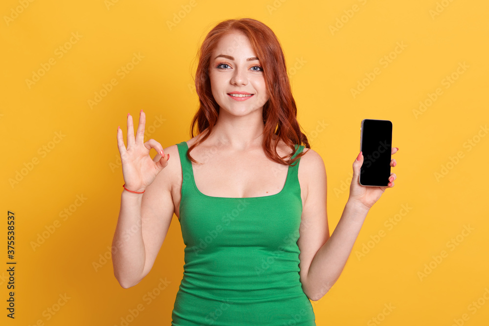 Young Caucasian woman wearing in green shirt showing ok sign on yellow background, holding mobile phone with blank screen, looks smiling directly at camera. Young Caucasian woman wearing in green shirt showing ok sign on yellow background, holding mobile phone with blank screen, looks smiling directly at camera.