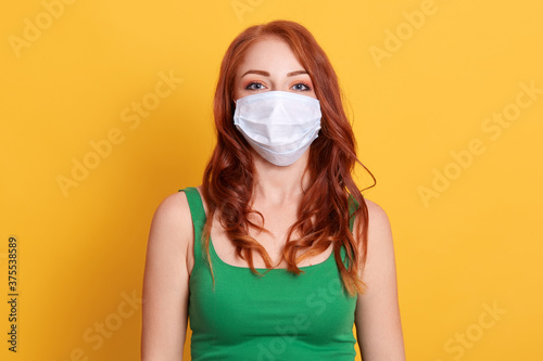 Young Caucasian woman wearing face mask to protect from COVID 19 isolated on yellow background, dresses green shirt, red haired lady looking at camera.