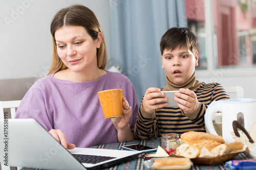 Portrait of young woman working at laptop and son using phone at table with tea indoors