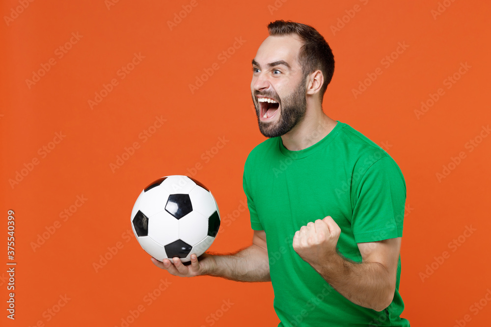 Happy man football fan in green t-shirt cheer up support favorite team with soccer ball clenching fist like winner looking aside isolated on orange background. People sport leisure lifestyle concept. Happy man football fan in green t-shirt cheer up support favorite team with soccer ball clenching fist like winner looking aside isolated on orange background. People sport leisure lifestyle concept.
