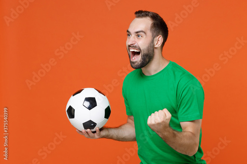 Happy man football fan in green t-shirt cheer up support favorite team with soccer ball clenching fist like winner looking aside isolated on orange background. People sport leisure lifestyle concept.