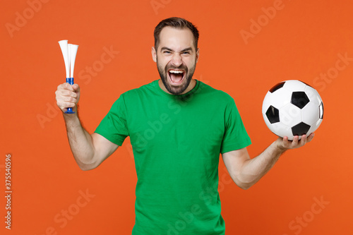 Cheerful young man football fan in basic green t-shirt cheer up support favorite team with soccer ball hold pipe isolated on orange background studio portrait. People sport leisure lifestyle concept.