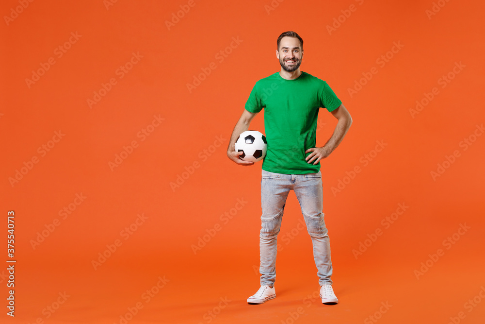 Full length portrait smiling young man football fan in basic green t-shirt cheer up support favorite team with soccer ball isolated on orange background studio. People sport leisure lifestyle concept.