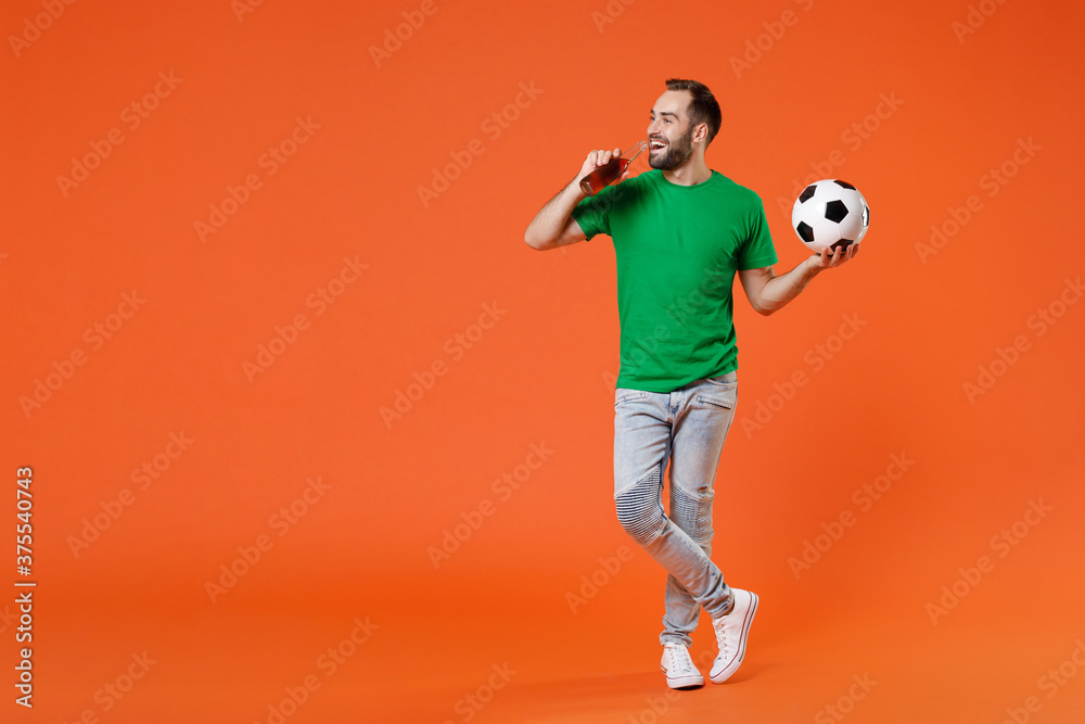 Full length portrait cheerful young man football fan in green t-shirt cheer up support favorite team with soccer ball hold beer bottle isolated on orange background. People sport leisure concept.