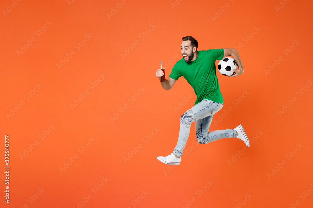 Full length portrait screaming man football fan in green t-shirt cheer up support favorite team with soccer ball hold beer bottle jumping isolated on orange background. People sport leisure concept.