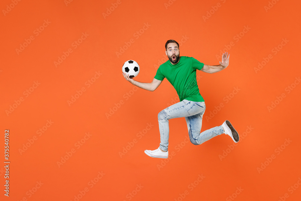 Full length portrait of shocked man football fan in green t-shirt cheer up support favorite team with soccer ball jumping spreading hands isolated on orange background. People sport leisure concept.