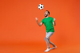 Full length portrait excited young man football fan in green t-shirt cheer up support favorite team throwing soccer ball isolated on orange background studio. People sport leisure lifestyle concept.
